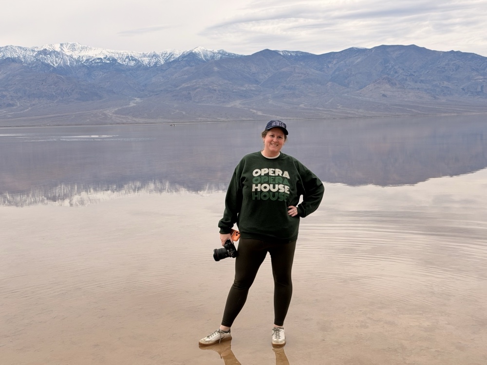 Heather Pierce standing in shallow water on a salt flat with her camera, snow-topped mountains reflected behind her